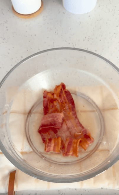 Cooked bacon strips sit in a clear glass bowl, which is placed on a light-colored cloth on a speckled countertop.