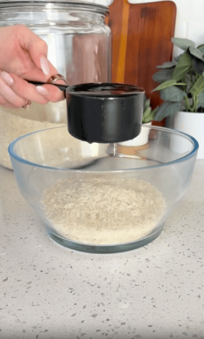 A hand pours a black measuring cup of rice into a clear glass bowl, with a jar of rice and potted plant in the background on a kitchen counter.