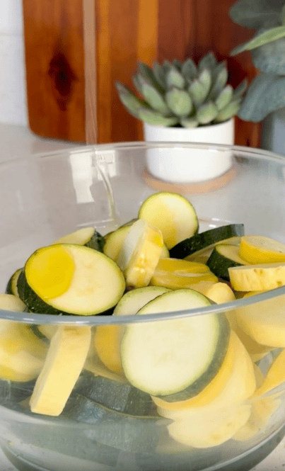 A glass bowl filled with sliced yellow and green zucchini sits on a counter, with a potted succulent plant visible in the background.