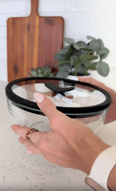 A hand holds a glass bowl with a black-sealed lid in a kitchen. In the background, there’s a wooden cutting board, a small potted plant, and a white countertop.