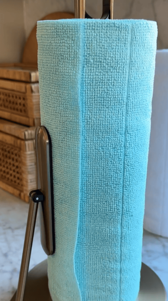A teal reusable cloth towel roll on a bronze paper towel holder, with woven baskets and a white item in the background on a marble countertop.
