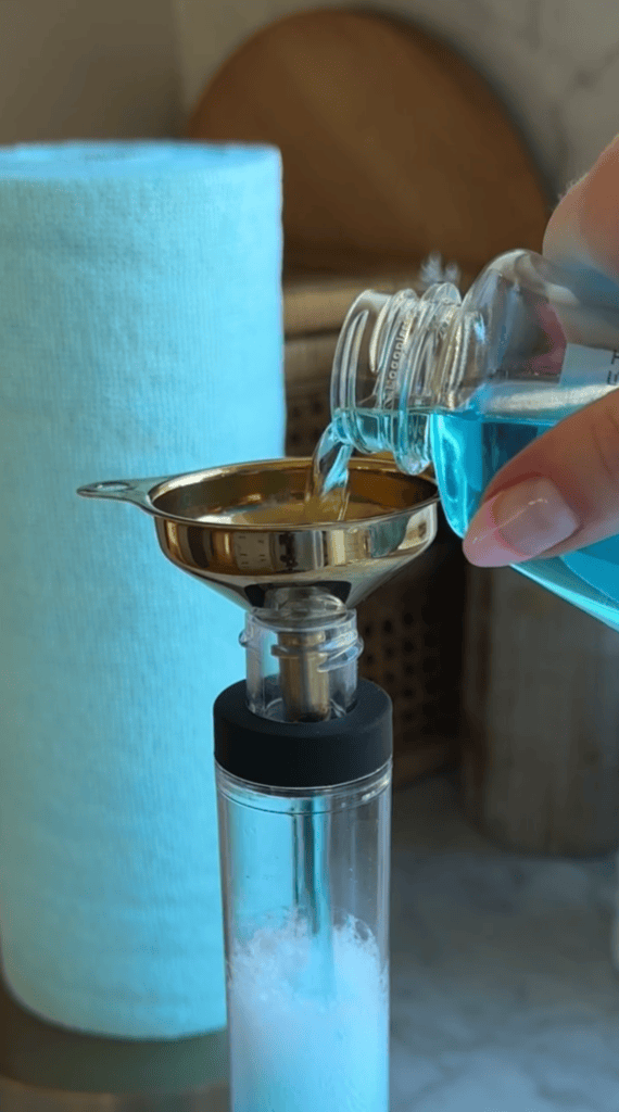 A hand pours blue liquid from a bottle into a gold funnel atop a clear container, with a roll of light blue paper towels in the background.
