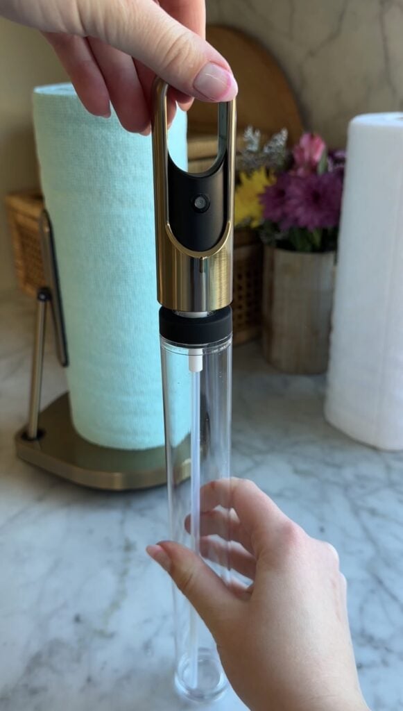 A person holds a clear tube with one hand and inserts a gold and black dispenser top with the other. Paper towels and flowers are in the background on a marble countertop.