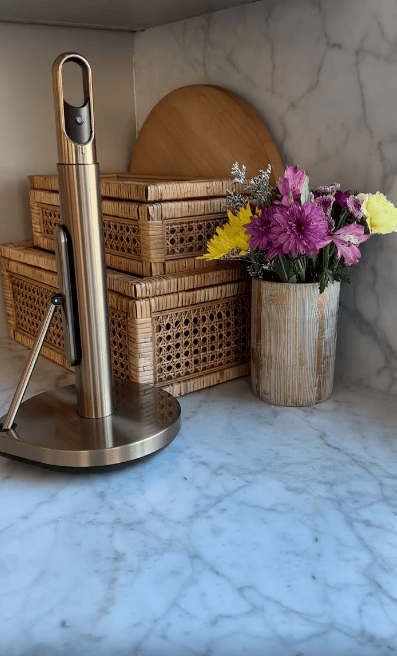 A marble countertop holds a paper towel holder, two woven boxes stacked, a round wooden board, and a bamboo-style vase with yellow and purple flowers.