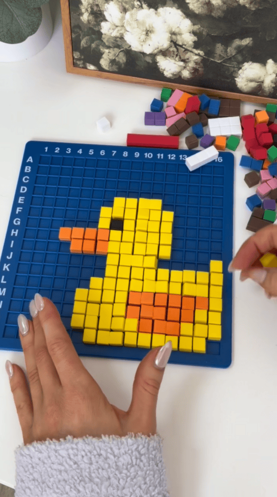 A person arranges colorful square blocks on a blue grid to form a pixel art image of a yellow duck with an orange beak and wing. More blocks are scattered nearby on the white surface.