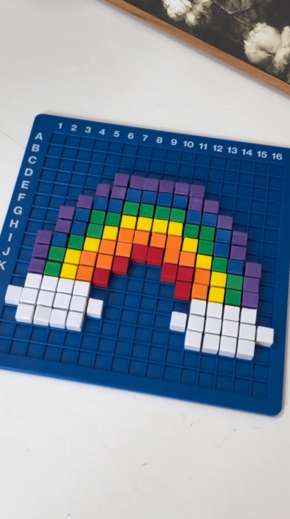 A blue grid features a pixel art rainbow with clouds, made from colorful square tiles arranged in rows and columns.