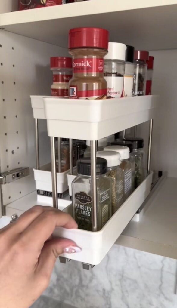 A hand pulls out a pull out spice rack from a kitchen cabinet, displaying organized spice containers such as parsley flakes and paprika.