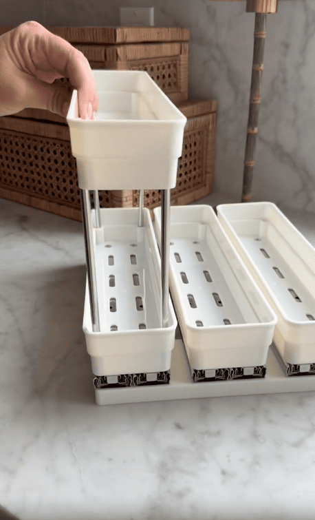 A hand lifts a white plastic storage bin with metal rods, reminiscent of a pull out spice rack, from a set of three bins on a countertop, with wicker boxes and a lamp in the background.