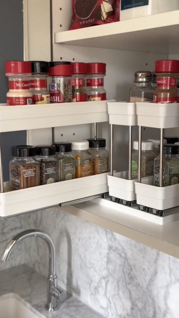 Two white tiered spice racks and a pull out spice rack are filled with various spice jars, all neatly organized on a kitchen countertop next to a sink with a marble backsplash. Some spices are also arranged on the shelf above the racks.