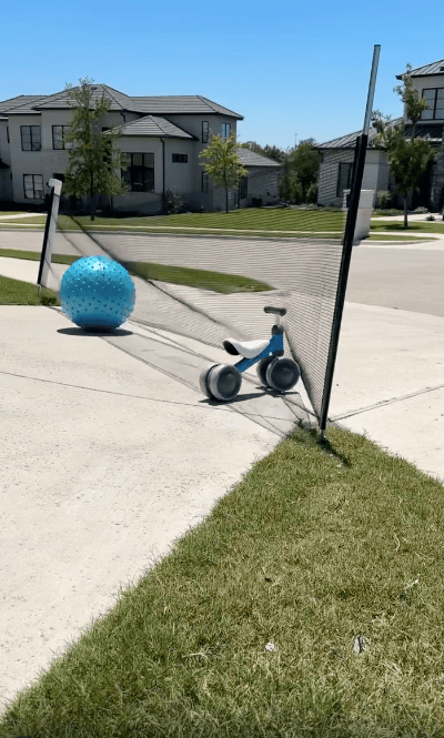 A blue exercise ball and a small blue tricycle are stuck in a black net set up on a suburban sidewalk, with houses and lawns visible in the background.
