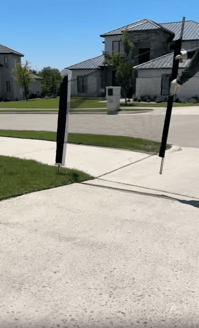 A small black and white net is set up across a driveway in a suburban neighborhood with grass and modern houses visible under a clear blue sky.