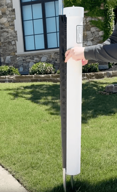 A person attaches a black and white protective cover to a vertical post on a grassy lawn, with a stone building and window in the background.