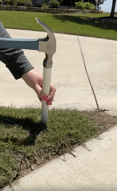 A person uses a hammer to drive a white plastic stake or pipe into the ground next to a concrete driveway on a sunny day.