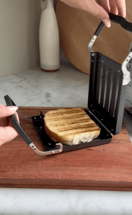 A person opens a black kitchen appliance, the Revolution Toastie Press, revealing a grilled sandwich with golden brown grill marks on a wooden cutting board—perfect for any toastie press review.