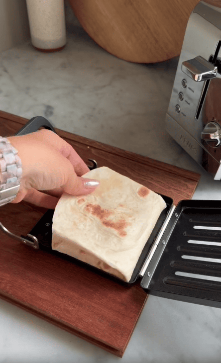 A hand places a folded tortilla wrap into a Revolution Toastie Press, a sleek kitchen appliance on a wooden countertop, with a toaster and cutting boards in the background.