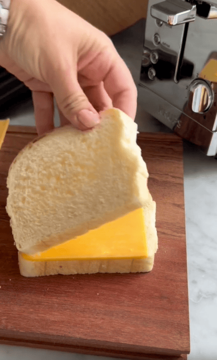 A hand lifts a slice of white bread to make a sandwich with cheddar cheese on another slice, set on a wooden board near the Revolution Toastie Press, the innovative kitchen appliance for perfect toasties.