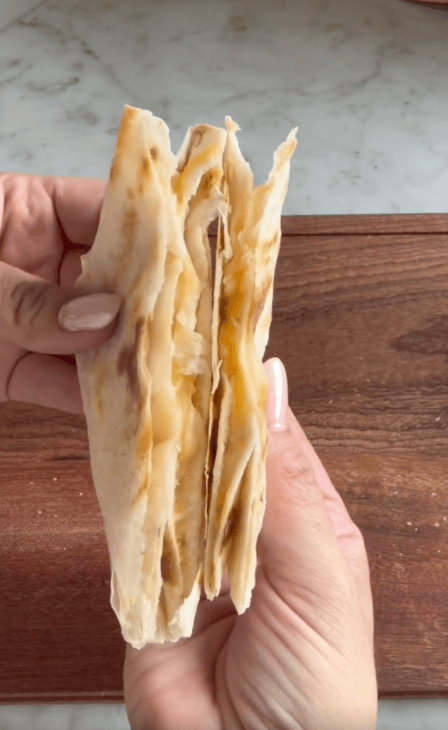 A hand holds a folded, flaky flatbread with melted cheese inside, over a wooden surface. The bread appears warm and freshly made, showing golden, crispy layers—perfect for testing in any Revolution Toastie Press review.