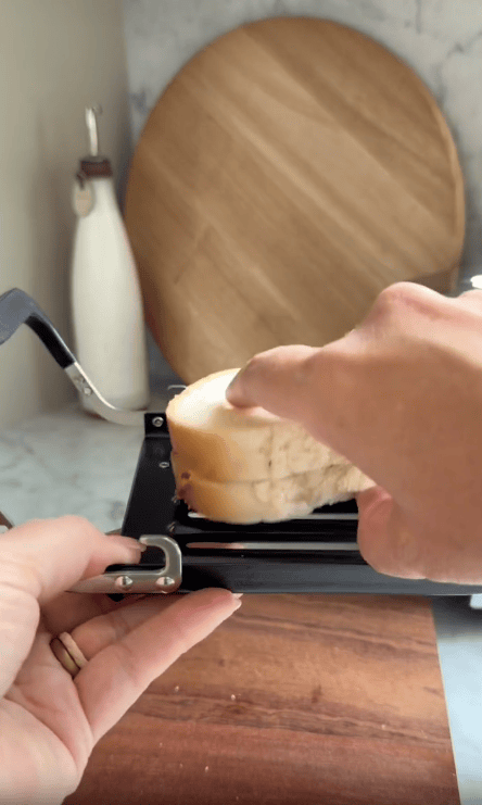 A hand slices a loaf of bread with a slicer on a wooden board, while an olive oil bottle and a Revolution Toastie Press kitchen appliance sit on a round cutting board in the background.