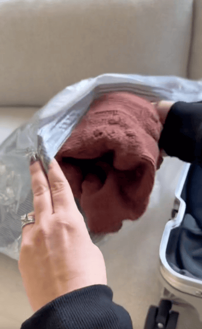 A person places a folded maroon towel into a large clear plastic bag, preparing items for packing into a suitcase that's partially visible on a beige couch.