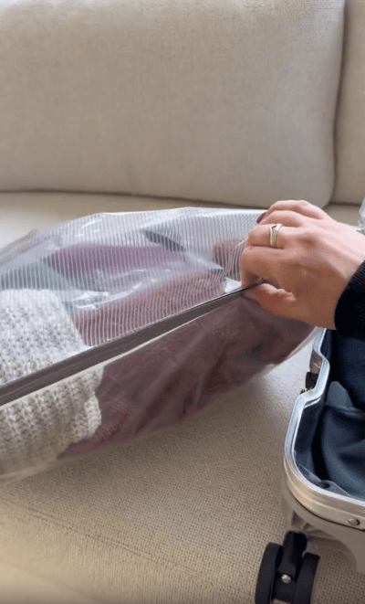 A person compresses clothes in a vacuum-sealed bag, pressing out air before packing it into a silver suitcase on a beige couch.