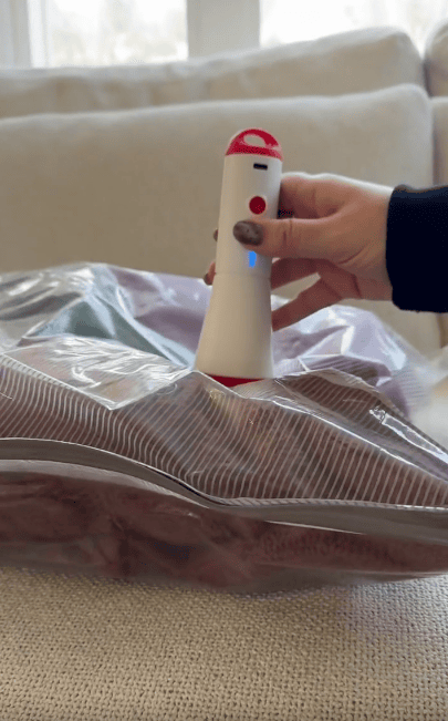 A hand uses a small, handheld vacuum sealer to remove air from a clear storage bag filled with folded clothes on a couch.