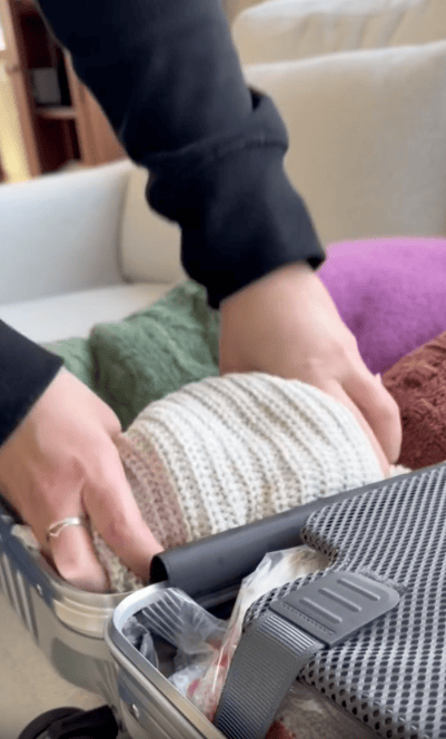 A person packs a suitcase with rolled clothing, including a white knit item, and a gray bag is visible in the front.