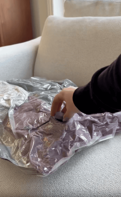 A hand is using a small device to seal or compress clothes inside a transparent vacuum storage bag on a light-colored couch.