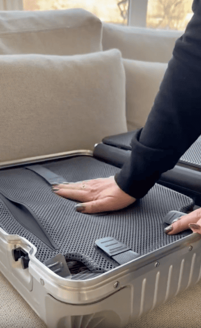 A person presses down on clothes packed in an open silver suitcase on a beige couch, securing them with inside straps.