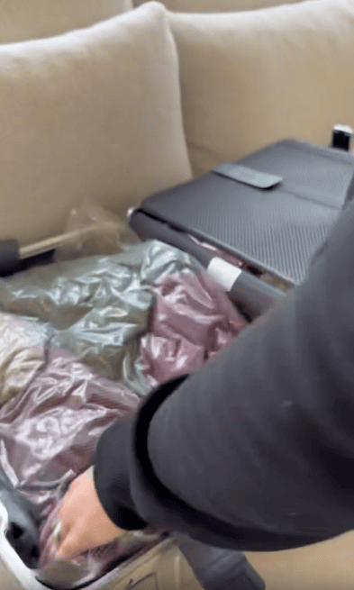 A person packs a suitcase with vacuum-sealed clothes on a beige couch. A briefcase or laptop bag is beside the suitcase.