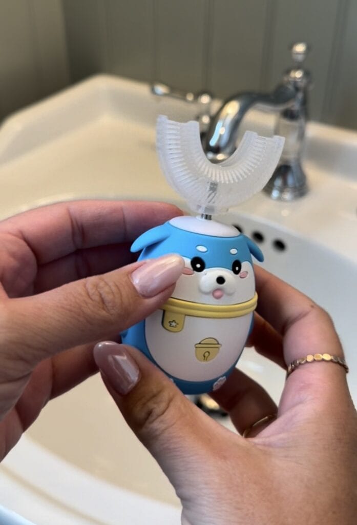 A person holds a blue and white otter-shaped automatic toothbrush with a U-shaped brush head, standing near a white bathroom sink and silver faucet.