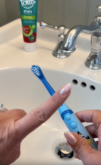 A hand points to a blue children's electric toothbrush over a white sink, with a tube of Tom's kids' toothpaste visible in the background near the faucet.