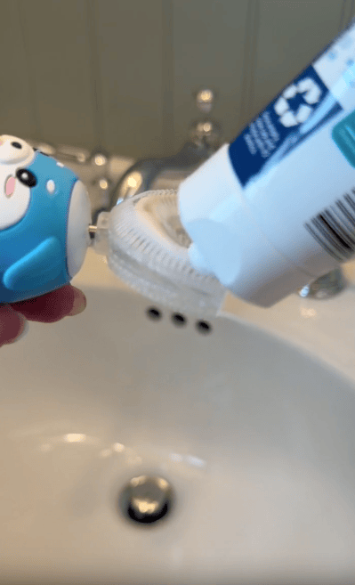 A person squeezes toothpaste onto a blue penguin-shaped toothbrush over a white sink, with a silver faucet and blurred background visible.