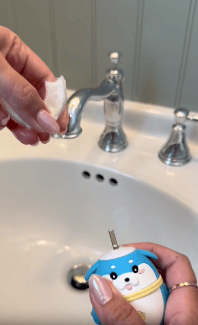 A person holds a blue animal-shaped contact lens case and a cotton pad near a white bathroom sink with a silver faucet.