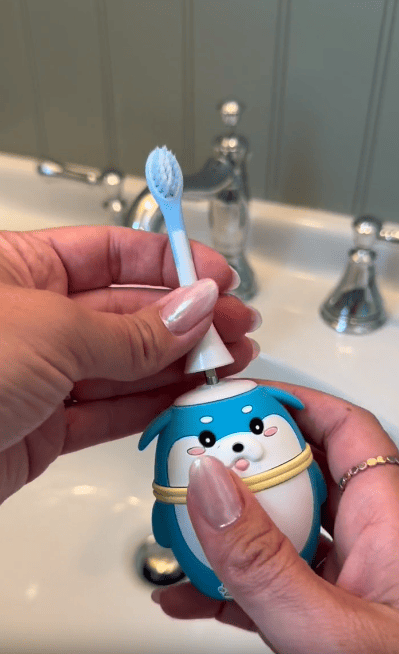 A person holds a blue, seal-shaped electric toothbrush and attaches a brush head above a bathroom sink with silver faucets.