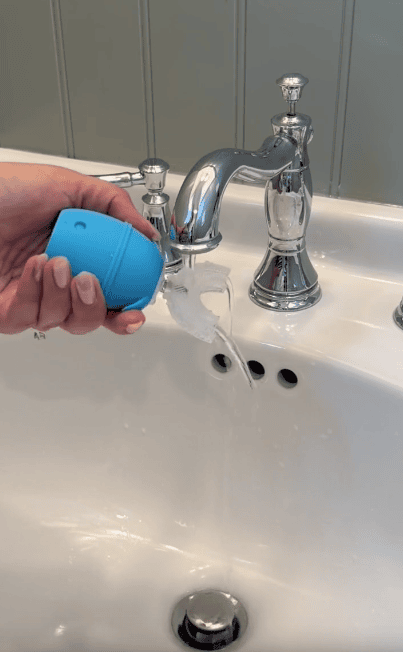 A hand holds a blue silicone device under a running bathroom faucet, altering the water flow to make it curve outward into the sink.