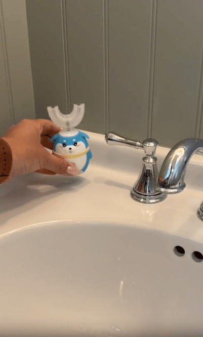 A hand holds a blue, shark-shaped automatic toothbrush with a U-shaped mouthpiece over a white sink next to a silver faucet.