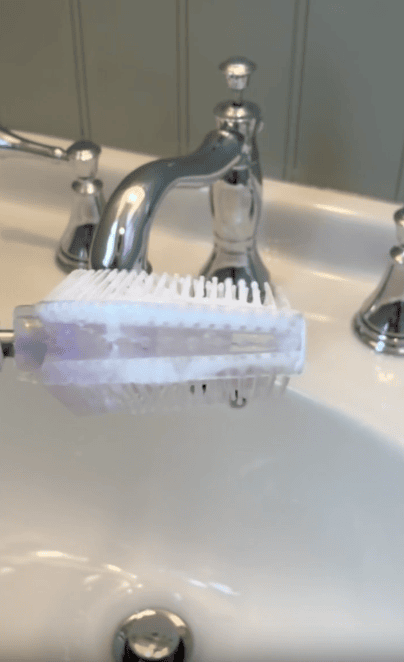 A white scrubbing brush with soap is held over a bathroom sink with chrome faucet and handles.