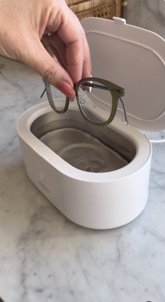 A hand lowers eyeglasses into a white ultrasonic cleaner filled with water, placed on a marble surface. The cleaner’s lid is open.