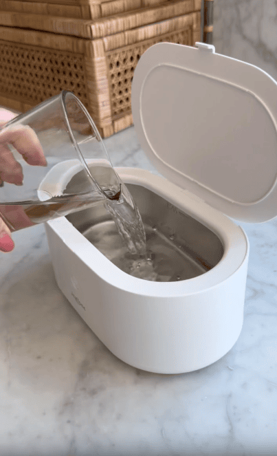 A person pours water from a glass into a white, oval-shaped ultrasonic cleaner with an open lid, sitting on a marble countertop.