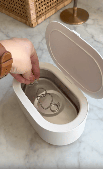 A hand lowers a ring into a white ultrasonic jewelry cleaner filled with water, where a bracelet and another ring are already submerged. The scene is set on a marble surface.