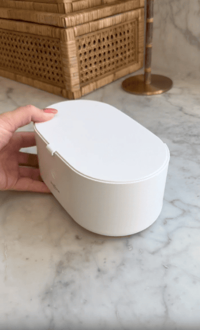 A hand holds a white, oval-shaped container with a hinged lid, set on a marble countertop with a wicker box and a lamp in the background.