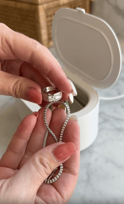 A person holds a silver ring and bracelet with manicured hands over a white jewelry cleaning machine on a marble surface.