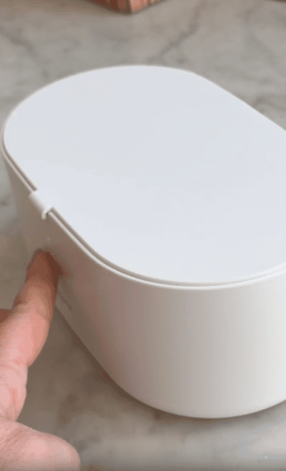 A hand presses a button on the side of a closed, oval-shaped white box placed on a marble surface.