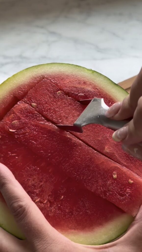 A person uses a Watermelon Slicer, a handy kitchen gadget, to cut rectangular slices into a halved watermelon, revealing the red flesh, seeds, and green rind—making life easier and mess-free.