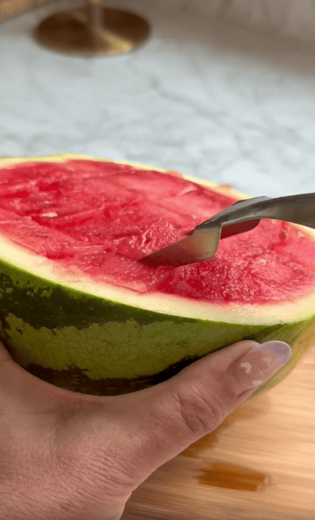 A hand holds a halved watermelon while a spoon scoops out the red fruit inside, resting on a wooden surface—upgrade to a watermelon slicer to make life easier and enjoy every bite effortlessly.