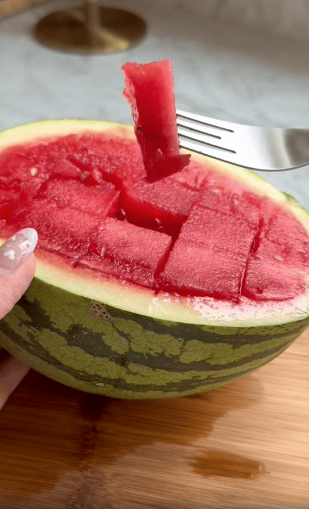 A hand holds half of a watermelon with its red flesh cut into cubes, while a fork lifts one cube. This clever kitchen tool makes life easier by neatly slicing the fruit, which sits invitingly on a wooden surface.