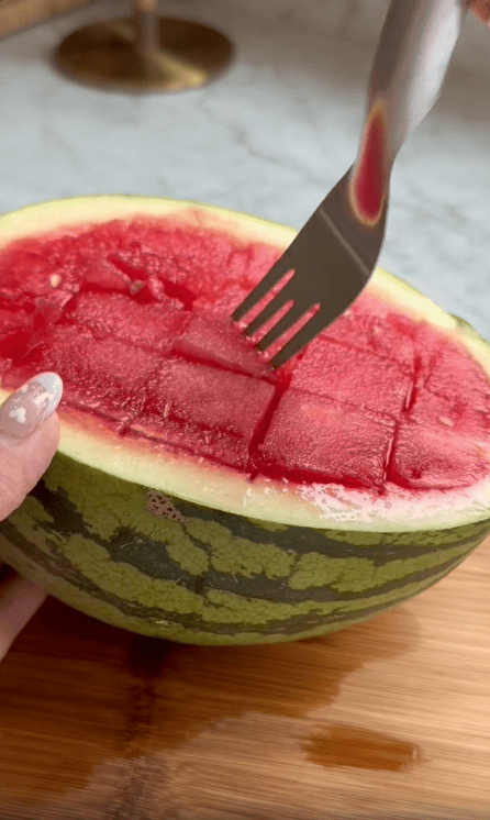 A hand holds half a watermelon with its red flesh cut into cubes, thanks to a handy watermelon slicer, while a fork is inserted into one of the cubes. The watermelon sits on a wooden surface, making life easier in the kitchen.