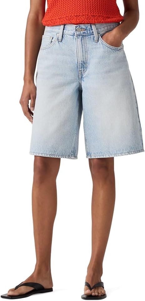 A person wearing light blue knee-length denim shorts, a sleeveless orange top, and black sandals, standing with one hand in their pocket.