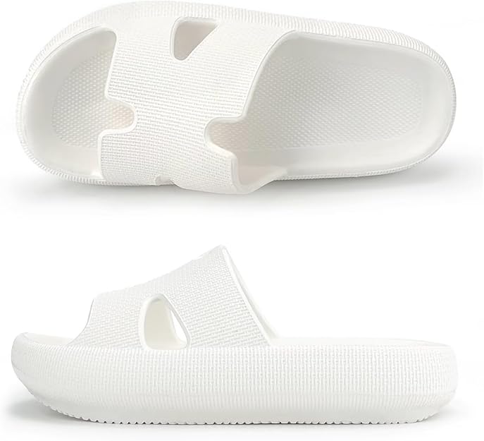 Two white, thick-soled slide sandals are shown. One sandal is placed on its side displaying the profile, while the other is positioned to show the top view, revealing textured surfaces and cut-out side openings.