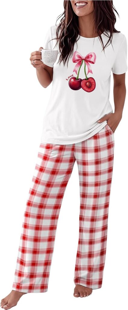 A smiling woman holds a white mug, wearing a white T-shirt with cherries and a bow graphic, and red-and-white plaid pajama pants.